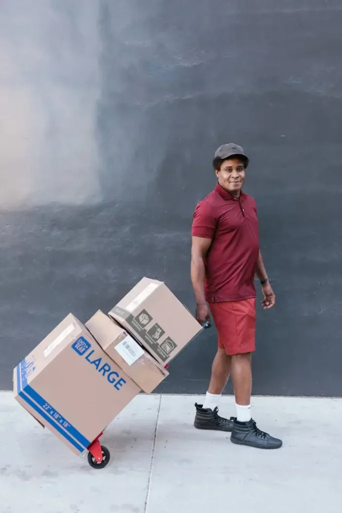 Smiling courier in red uniform transporting cardboard boxes outdoors using a hand truck.